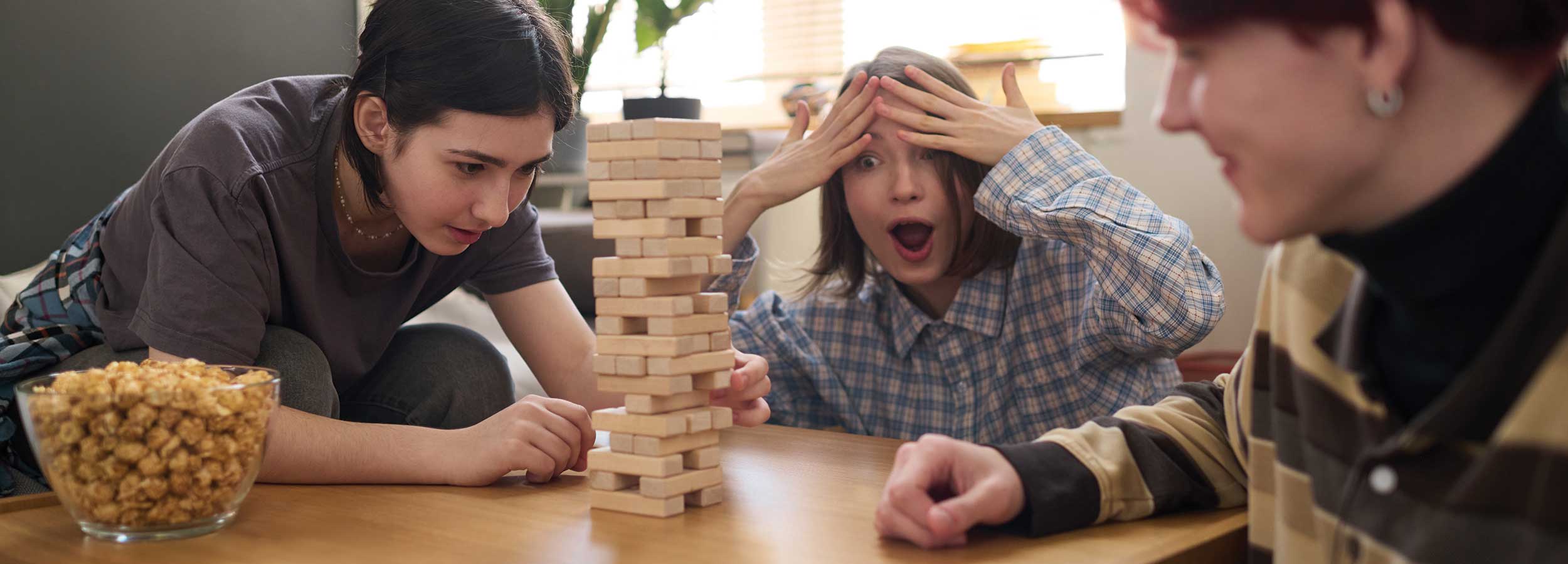 Three teenagers playing Jenga, with one carefully removing a block while another reacts with shock and hands on head, seated around a wooden table with a bowl of popcorn nearby.